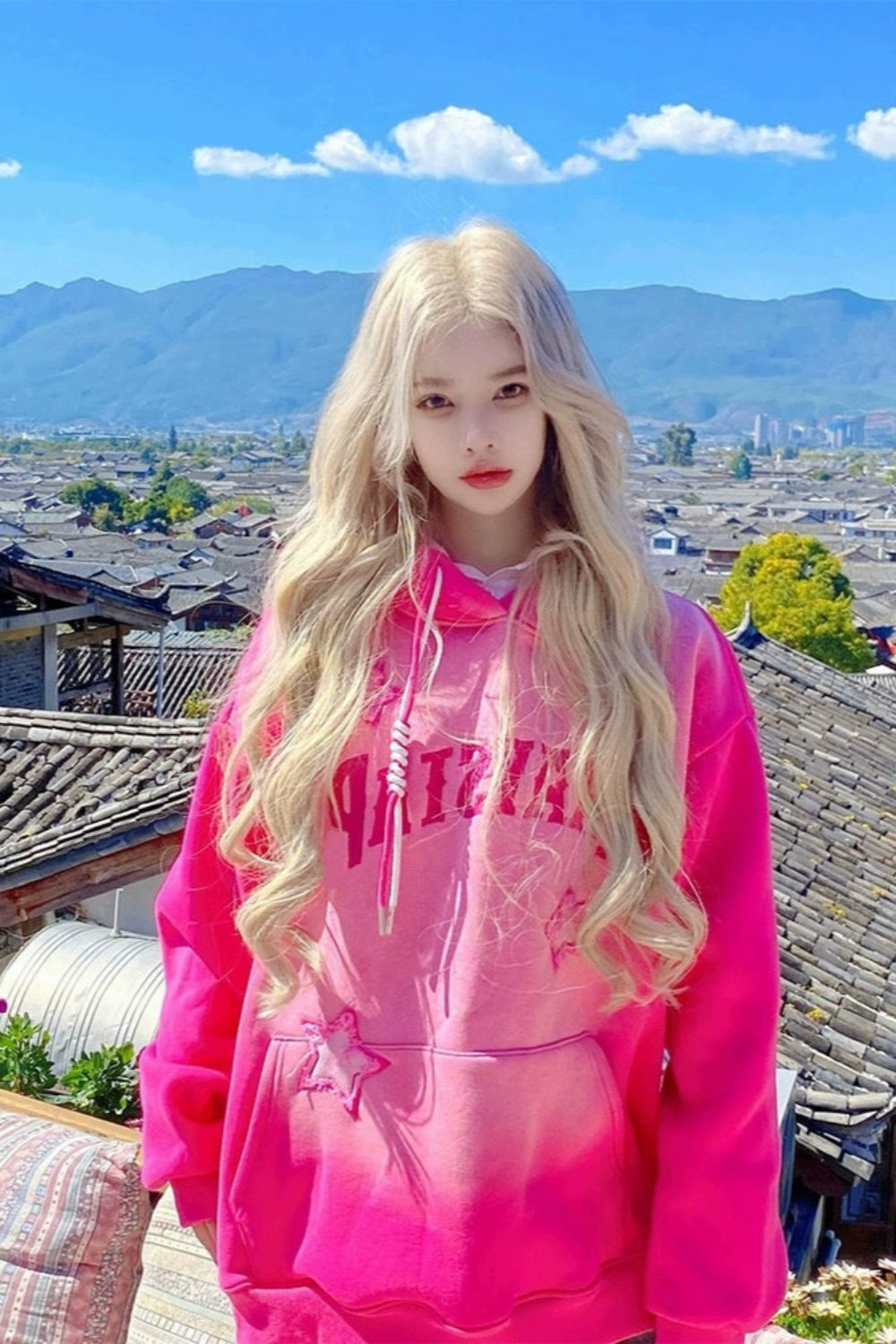 Person wearing a bright pink hoodie with a scenic background of mountains and buildings.